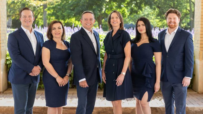 Six adults dressed in formal navy blue attire stand side by side outdoors, smiling and posing for a group photo with greenery and flowers in the background.